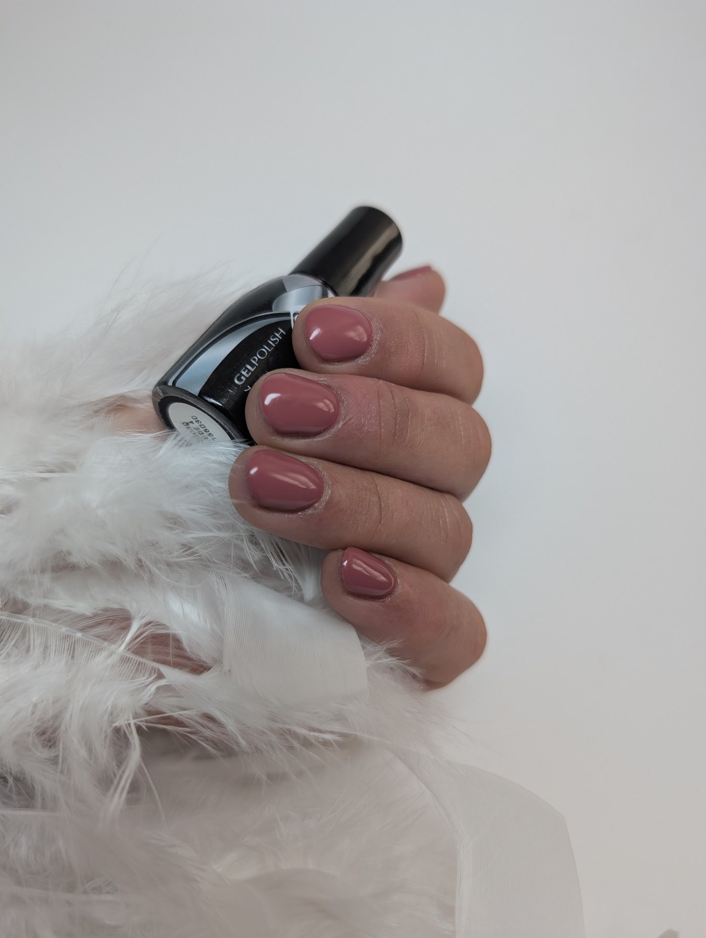 Korte, natuurlijke nagels in een warme mauve gellak naast een flesje gellak en witte veren