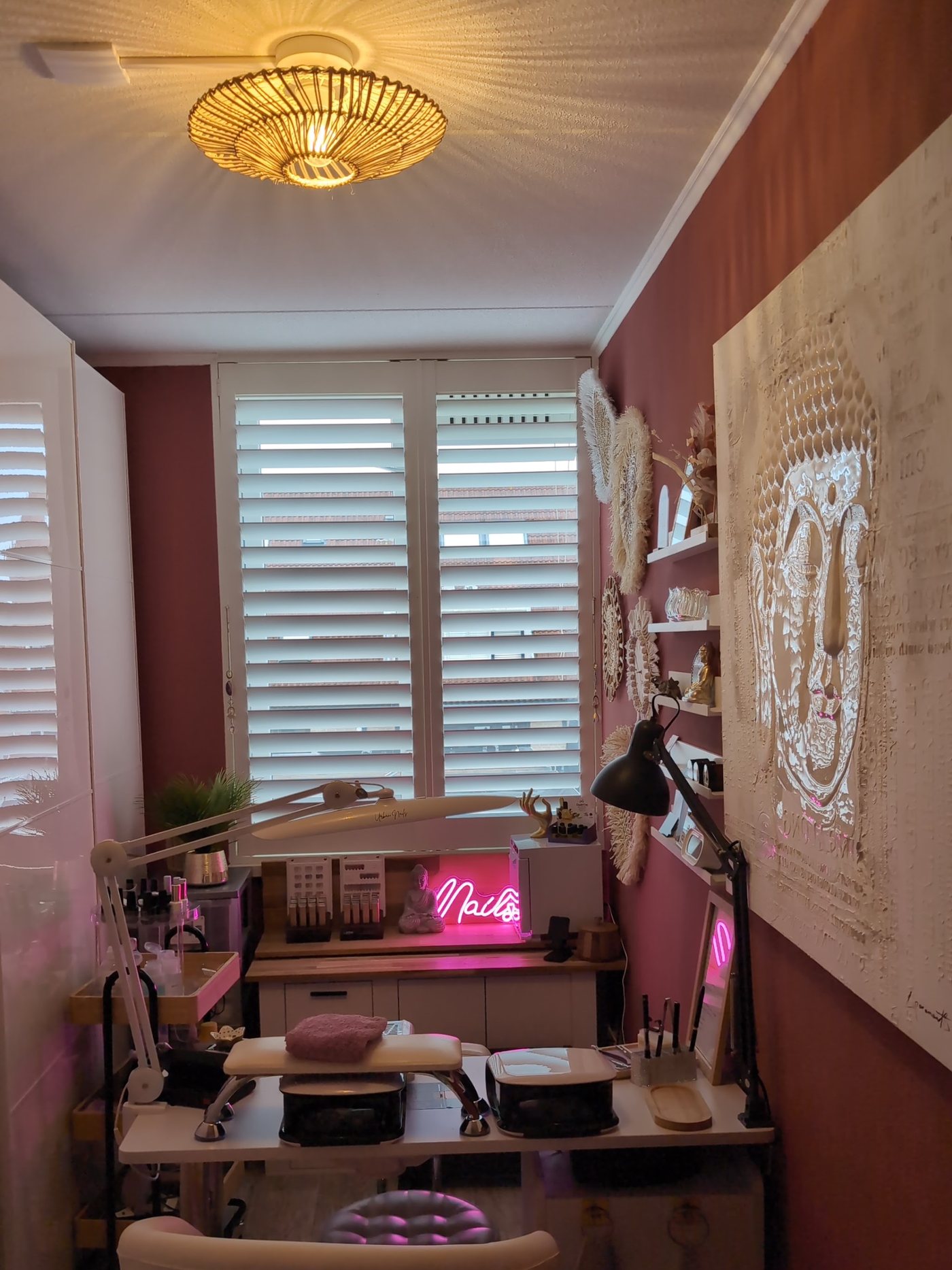 De studio vanaf de deur: rotan lamp, witte shutters, roze neon Nails en een groot Boeddha-doek op de terracotta muur